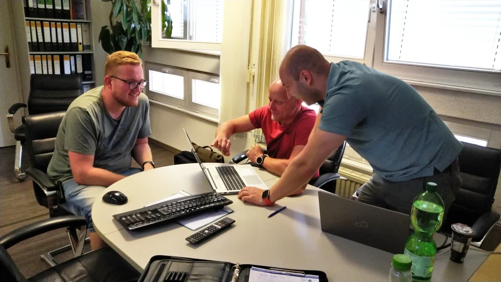 Auf dem Bild sieht man zwei Mitarbeitende von Holz Hess und einen Mitarbeitenden vom Mittelstand-Digital Zentrum Chemnitz. Alle beugen ich über einen Laptop und besprechen die Implementierung der Software-Lösung